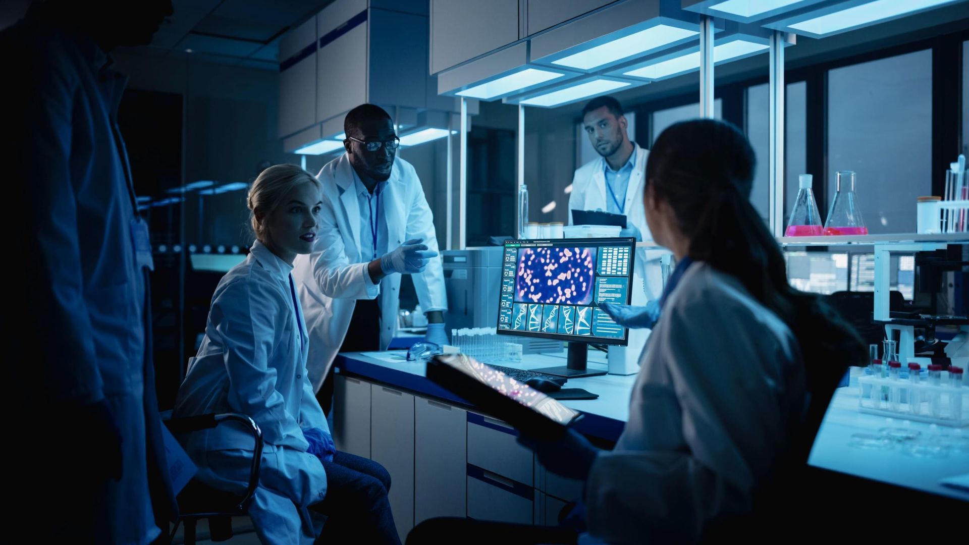 Scientists in blue-lit lab reviewing data on a computer monitor together