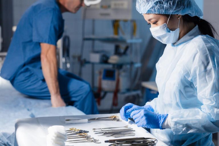 Healthcare workers in blue scrubs and protective gear preparing surgical instruments in an operating room