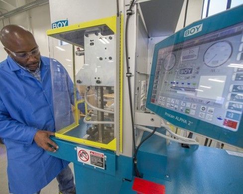 Man in blue lab coat operates BOY molding machine, examining controls at a factory.