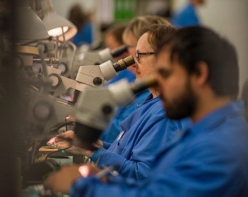 People in blue lab coats examining items through microscopes in a brightly lit lab.