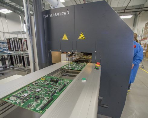 Circuit boards on conveyor belt entering a dark industrial soldering machine labeled 