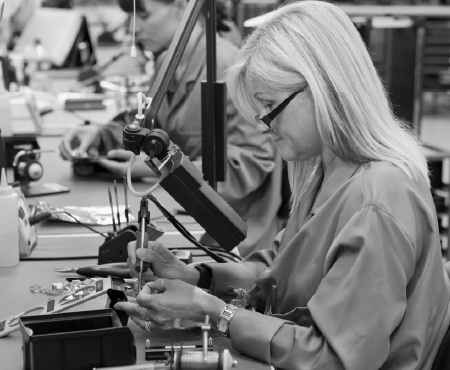 Two women in lab coats soldering at a workbench. One has gray hair and glasses.