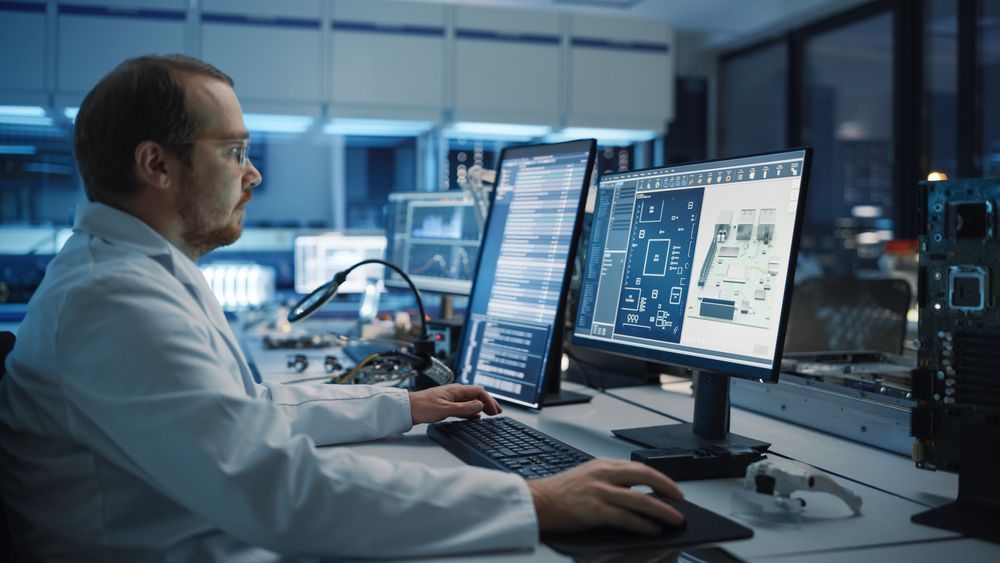 Man in lab coat working at computer monitors in a tech lab, focused on data and circuits.