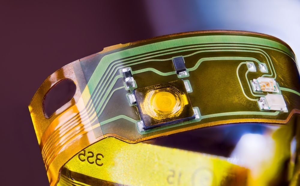 Flexible circuit board, amber-colored, curved, with electronic components and traces; close-up.