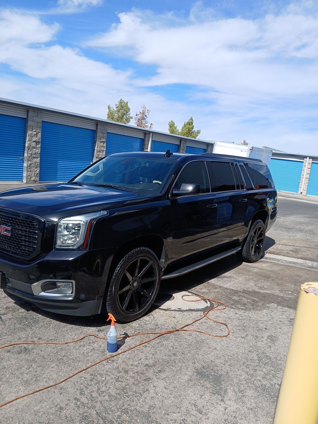 Black SUV parked by blue storage units on a sunny lot, with an orange cone in front.