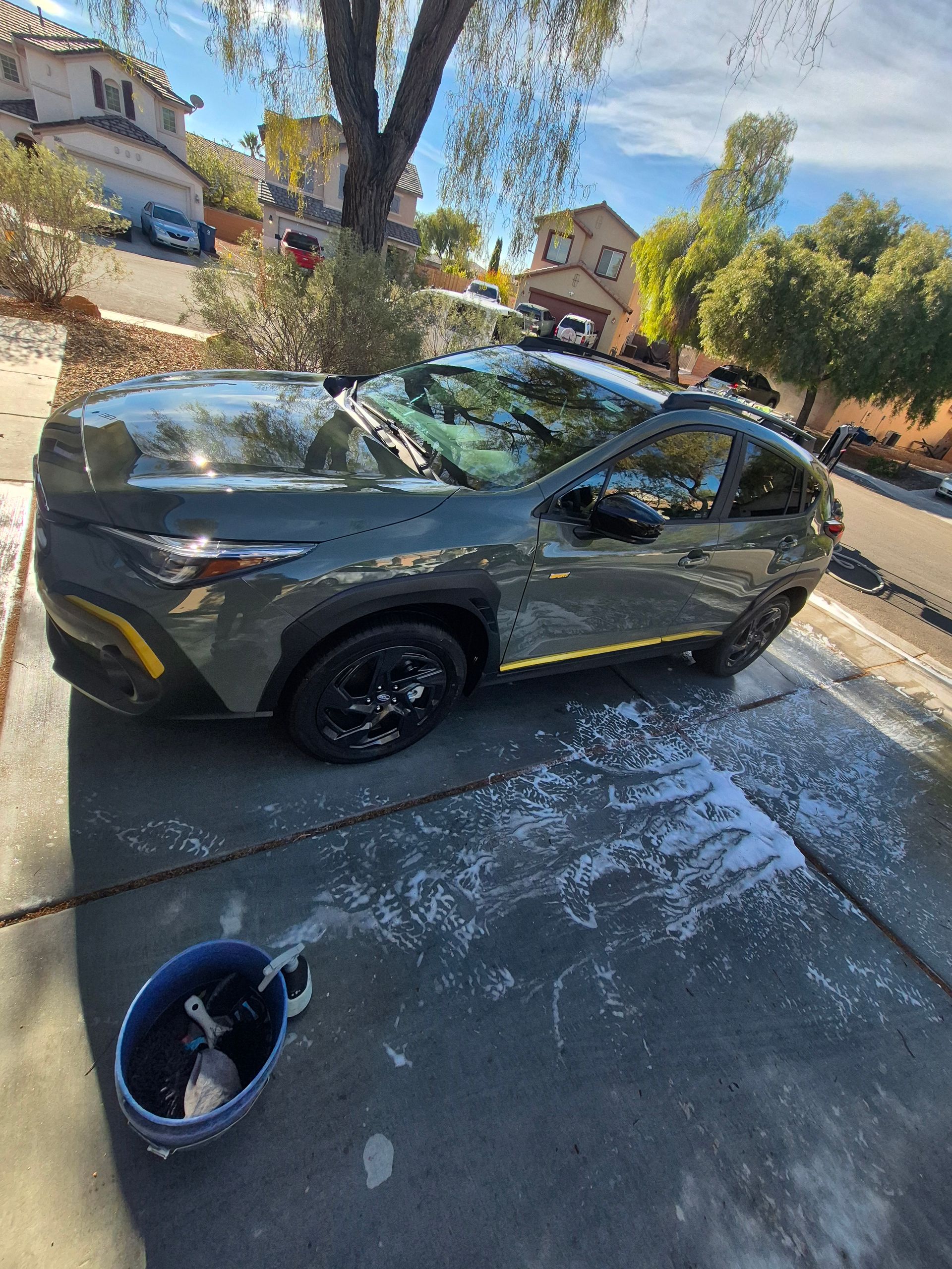 Dark green sedan being washed in a driveway, with a bucket in the foreground.