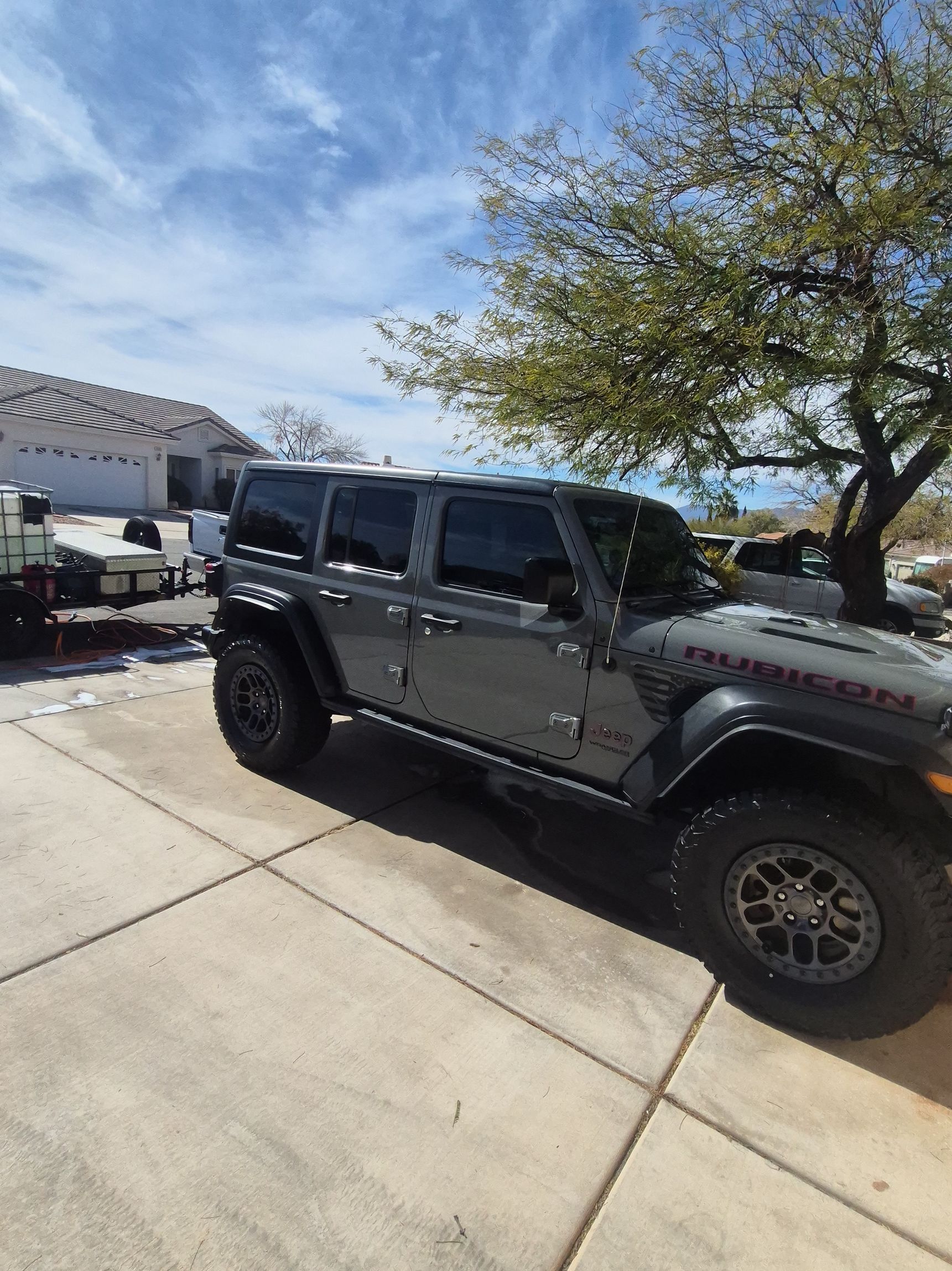 Gray Jeep Wrangler parked in a sunny driveway beside a house and tree.