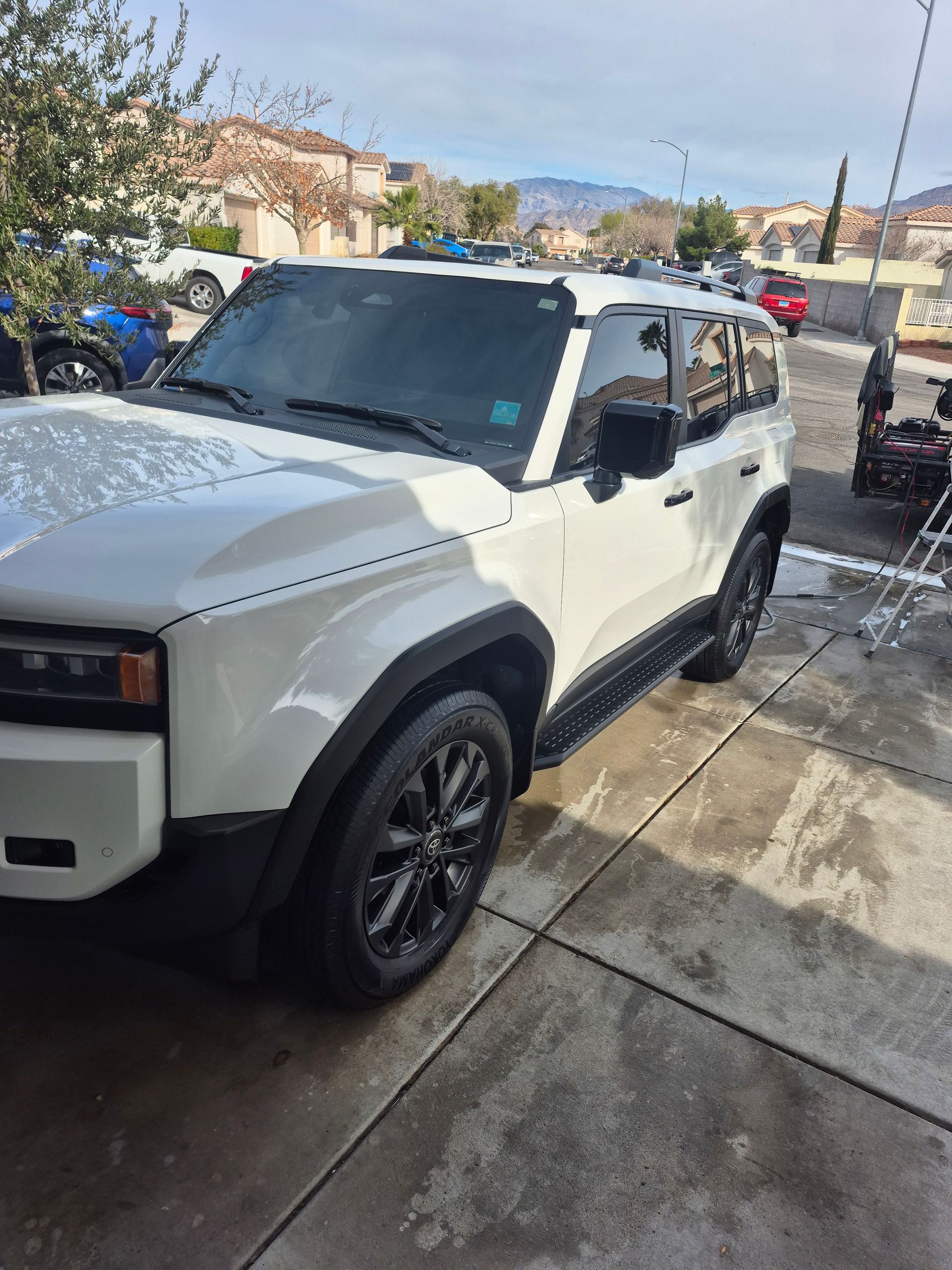 White off-road SUV parked on a wet driveway beside a house, with mountains in the background.