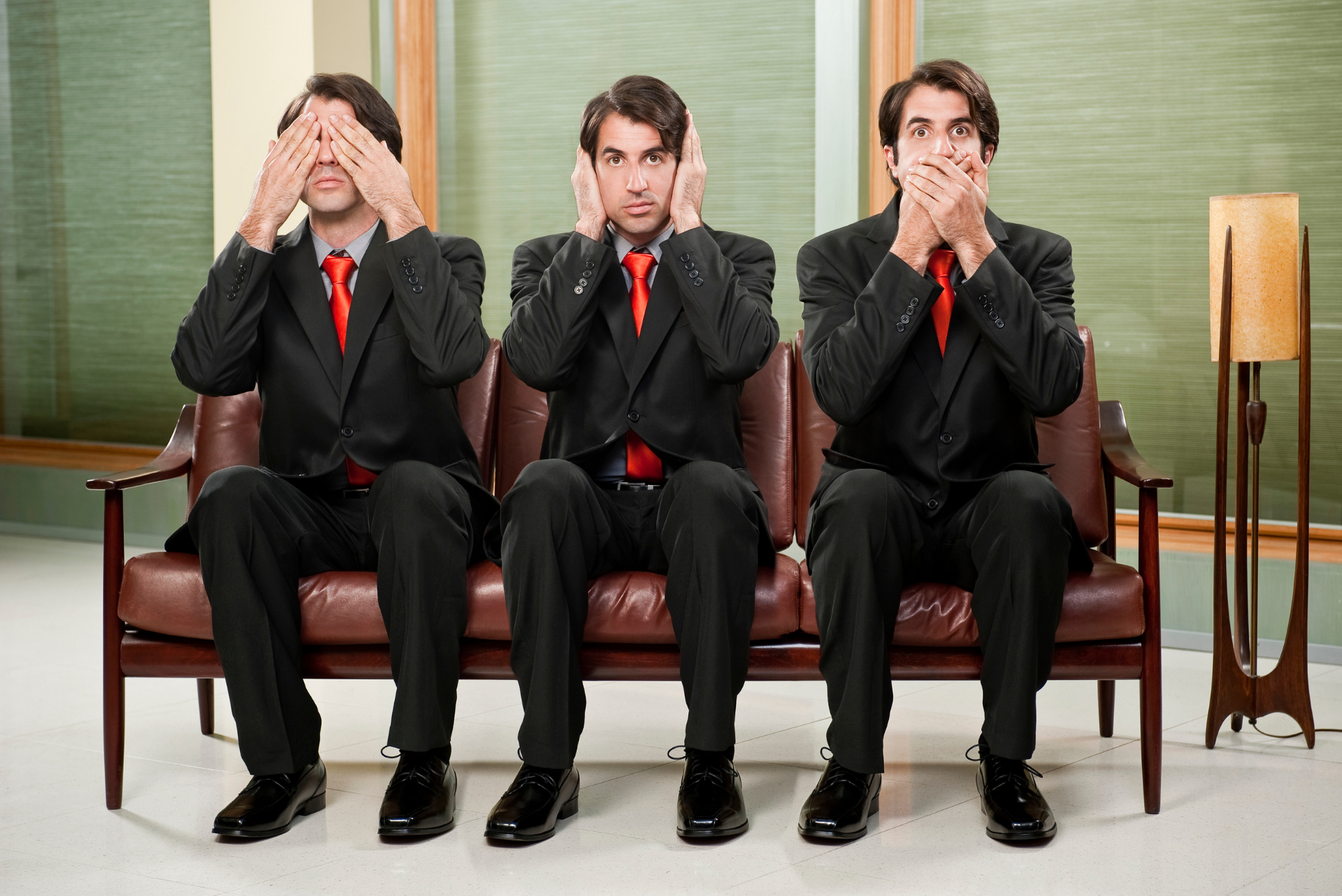 Three men in suits and ties are sitting on a couch covering their eyes , ears and mouths.