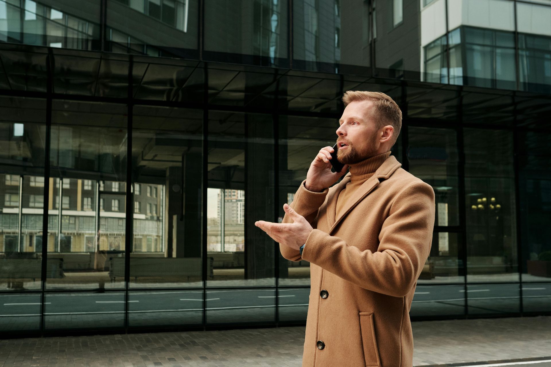 Man in tan coat talking on phone outside a modern glass building, gesturing with his hand.