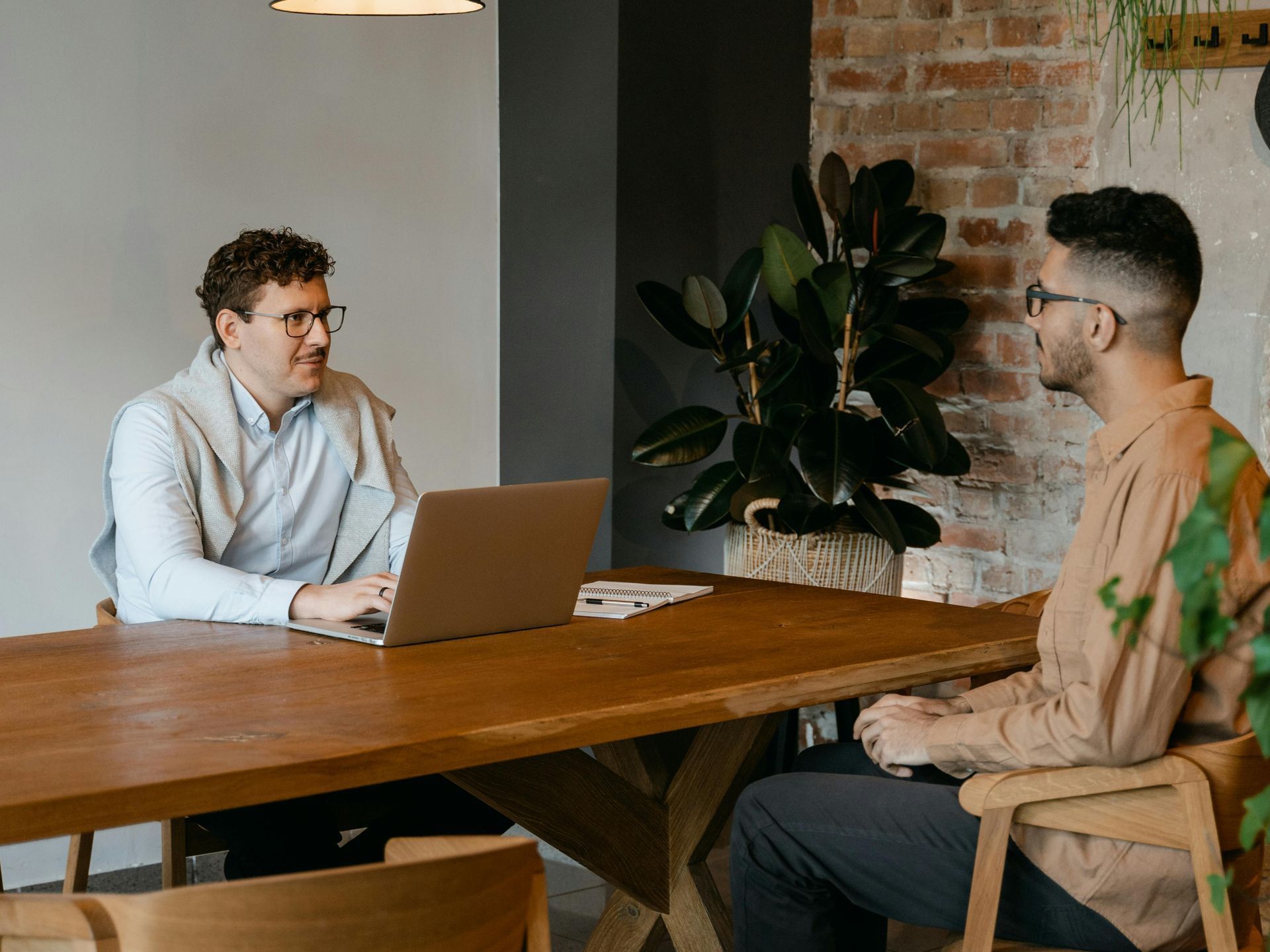 Two men are sitting at a table with a laptop and talking to each other.