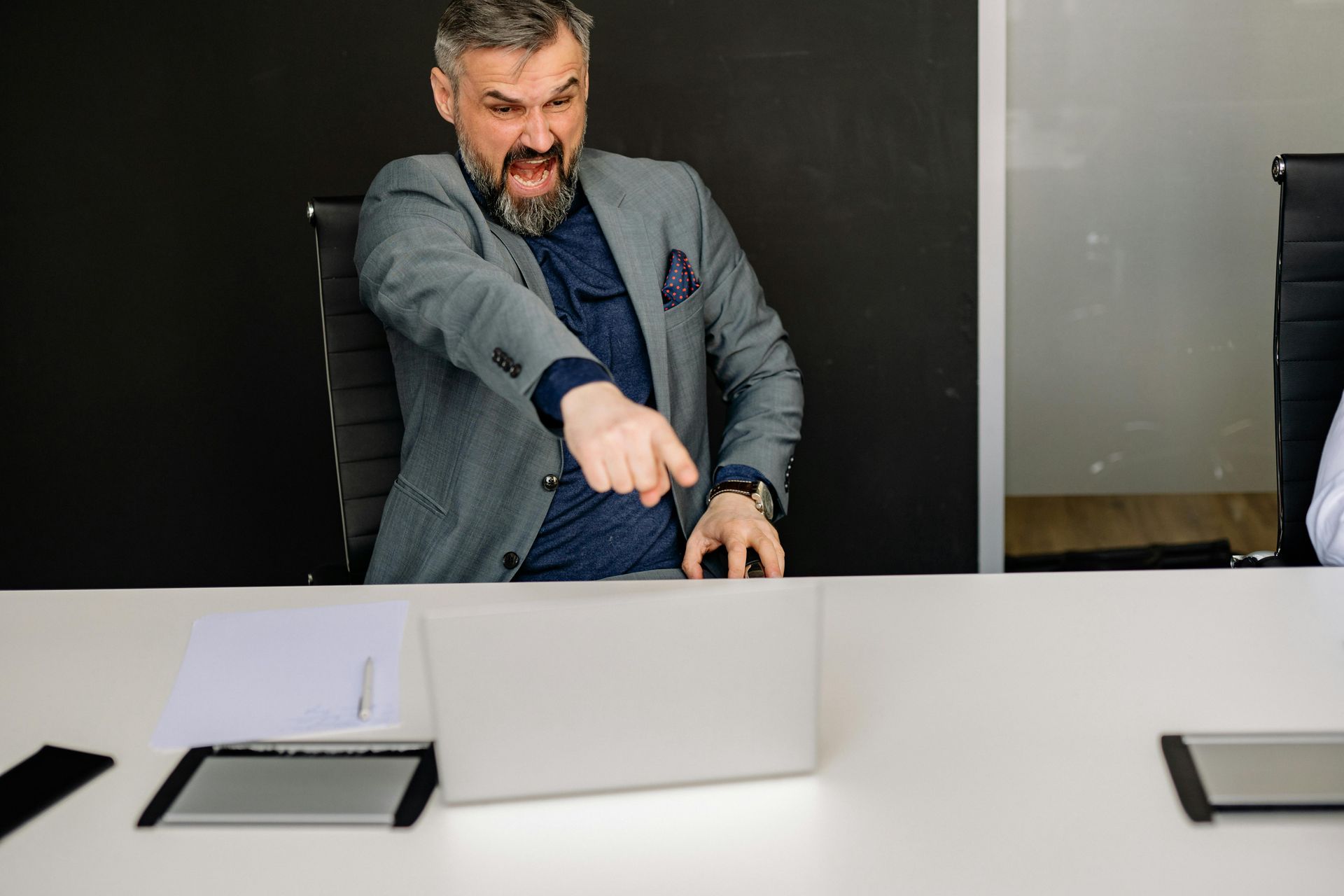An angry man is sitting at a table with a laptop and pointing.