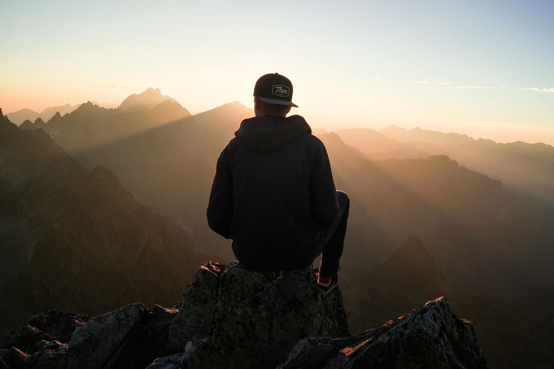 A man is sitting on top of a mountain looking at the sunset.