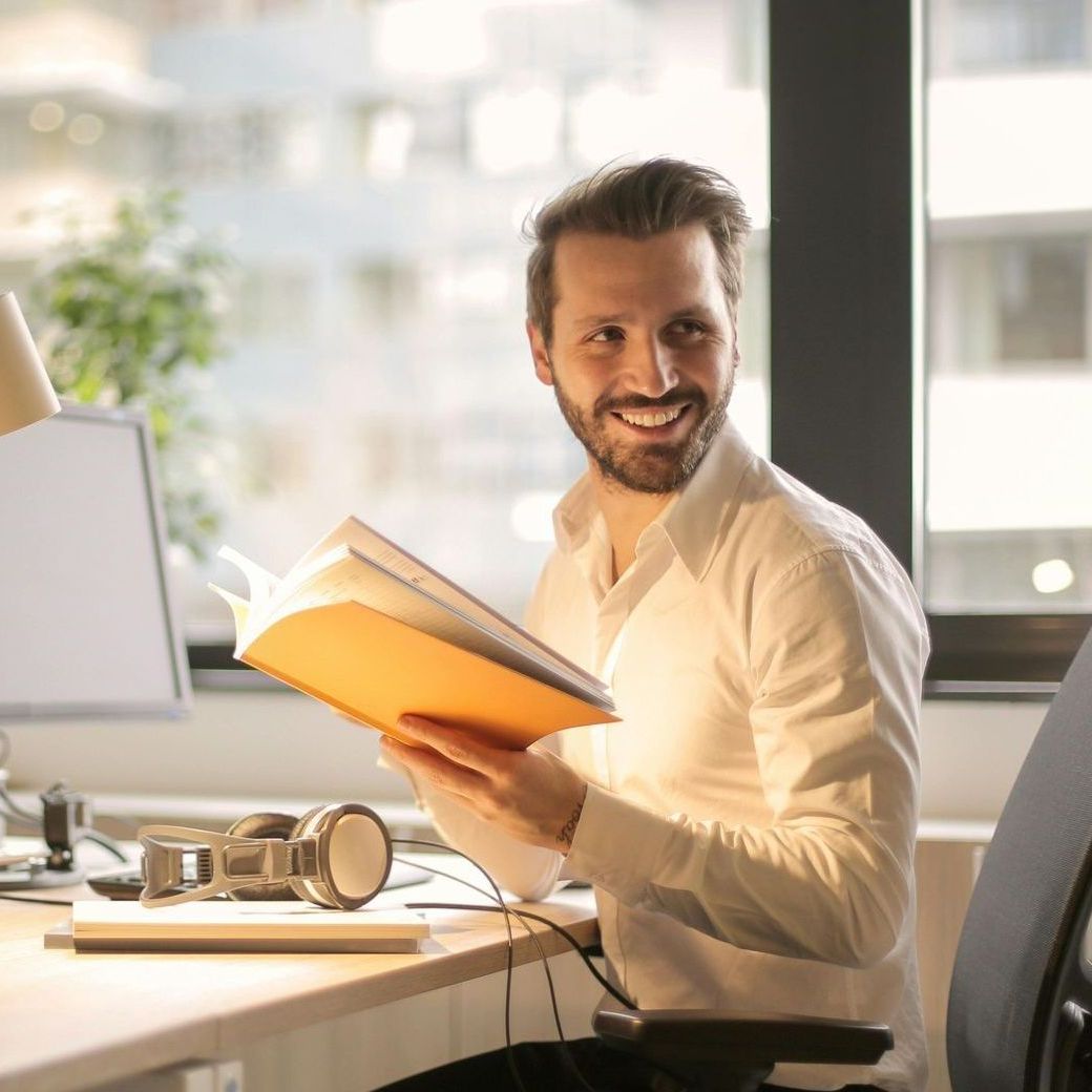 A man is sitting at a desk holding a book and smiling.