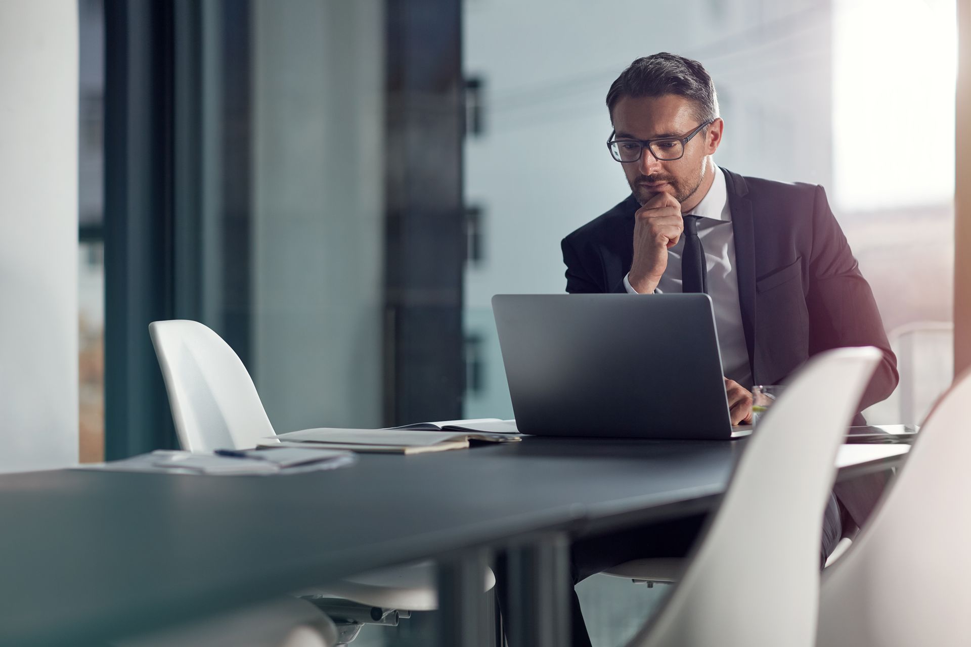 A professional in a suit and glasses sits at a table in a modern office, thoughtfully looking at a laptop screen.
