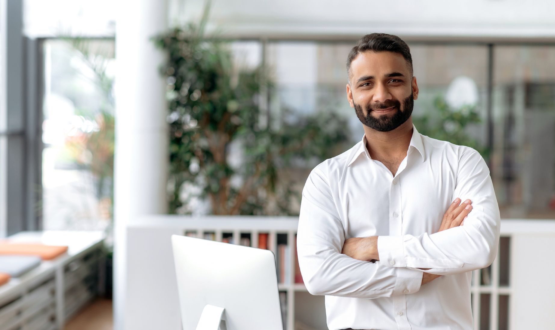 Man with a beard standing in front of a computer in an office with his arms crossed