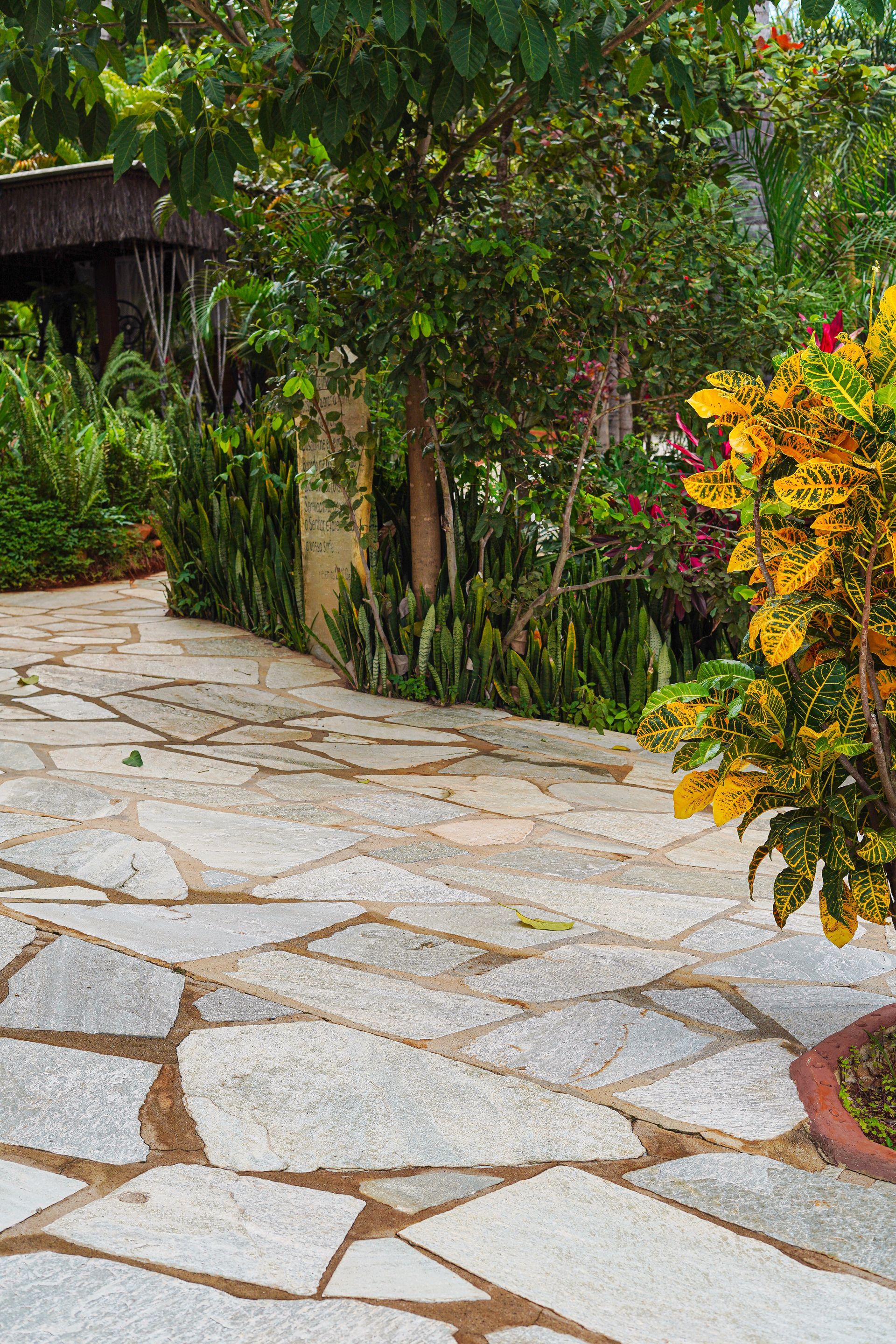 Pedra Lajão natural aplicada em pisos e caminhos externos, com grandes peças irregulares em tons de cinza, verde e bege integradas ao paisagismo tropical.