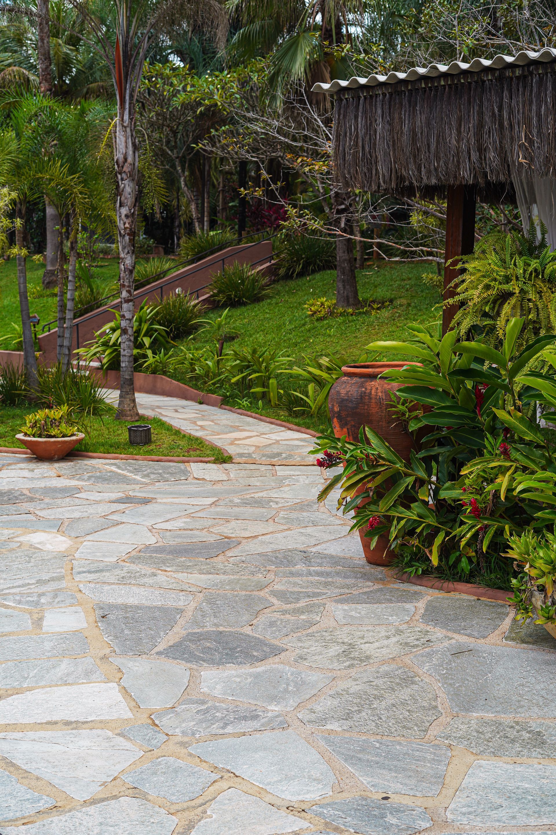 Pedra Lajão natural aplicada em pisos e caminhos externos, com grandes peças irregulares em tons de cinza, verde e bege integradas ao paisagismo tropical.