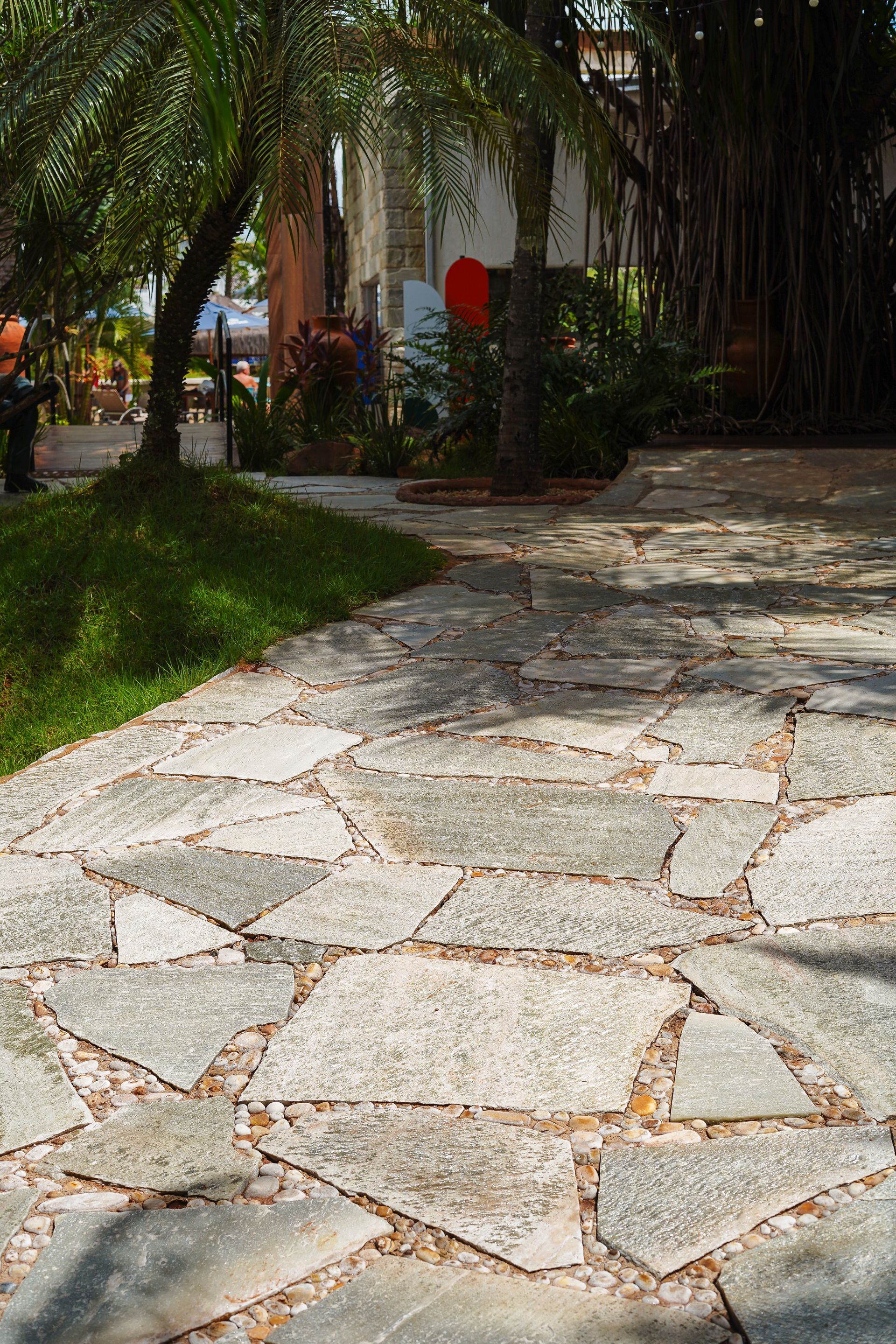 Pedra Lajão natural aplicada em pisos e caminhos externos, com grandes peças irregulares em tons de cinza, verde e bege integradas ao paisagismo tropical.