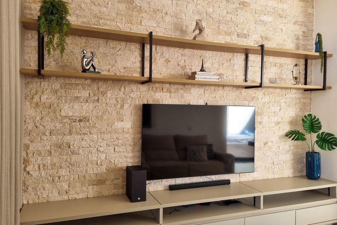 Sala de estar moderna com TV fixada na parede, prateleiras de madeira e vasos de plantas em uma parede com travertino nacional.
