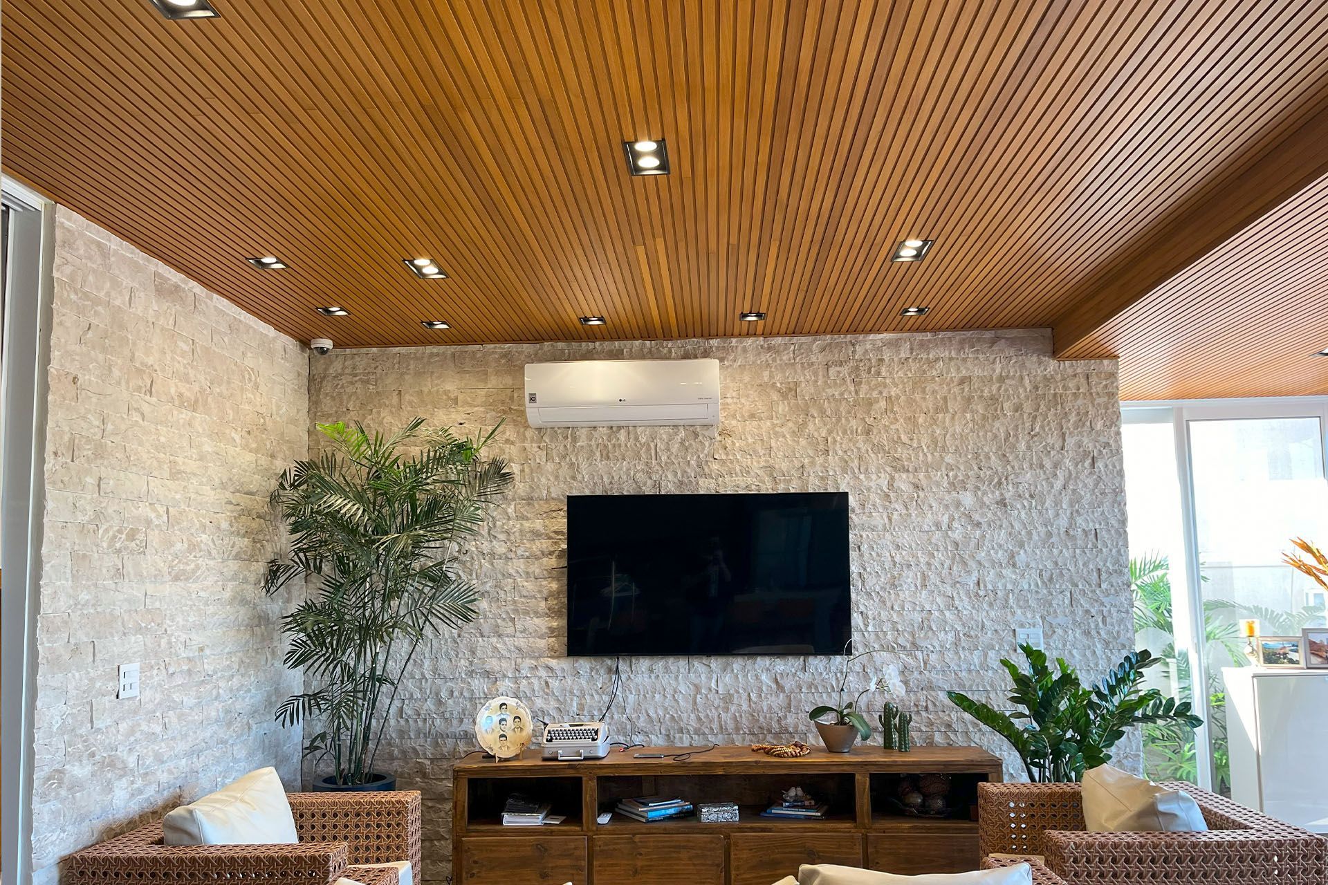Sala de estar moderna com teto de madeira, parede de pedra travertino nacional, TV fixada na parede e plantas de interior.