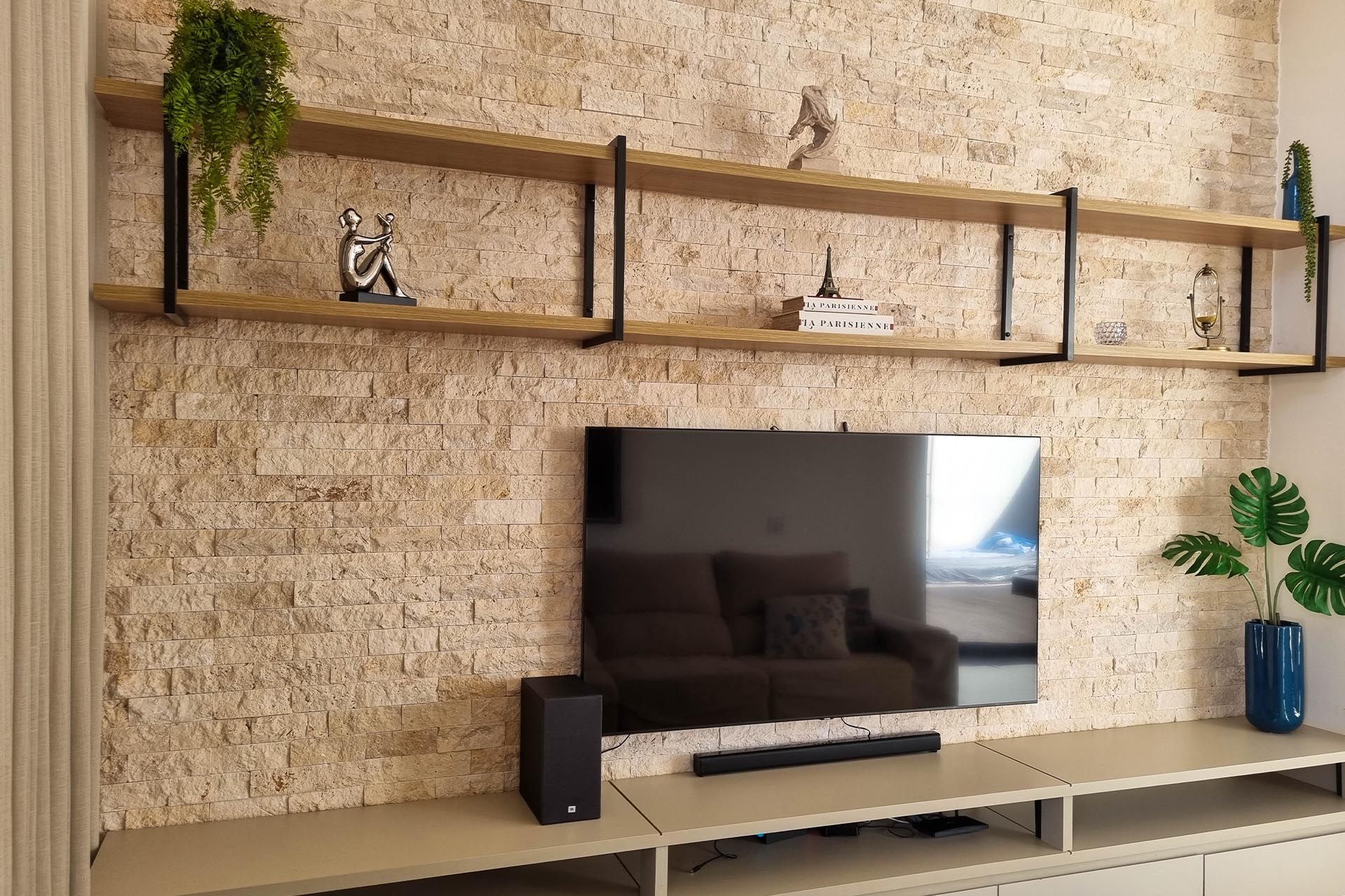 Sala de estar moderna com parede revestida de travertino nacional rockface, decorada com prateleiras flutuantes, TV, vasos de plantas e console de madeira clara.