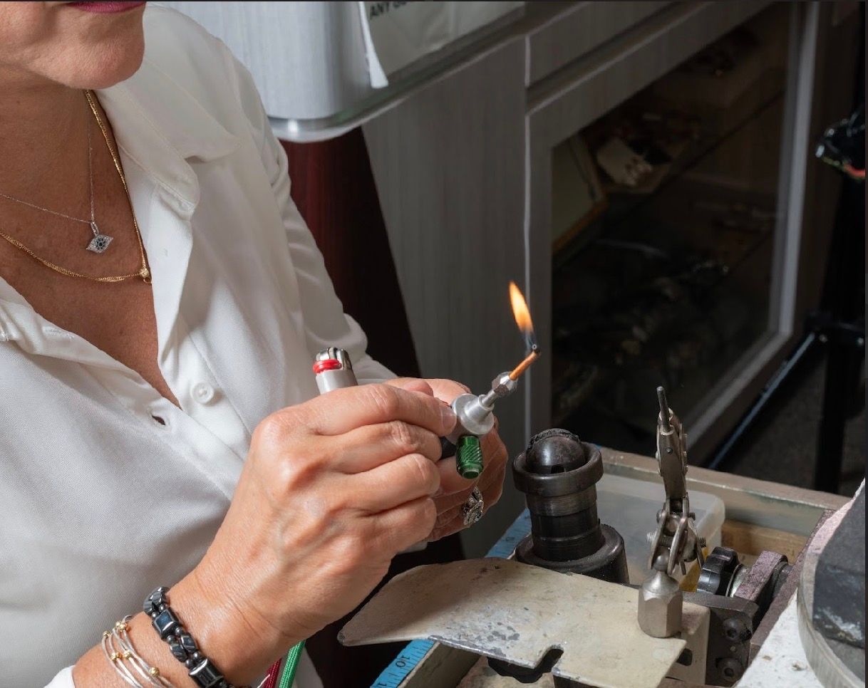 Person using a torch to heat a small object at a jeweler’s workbench near a window.