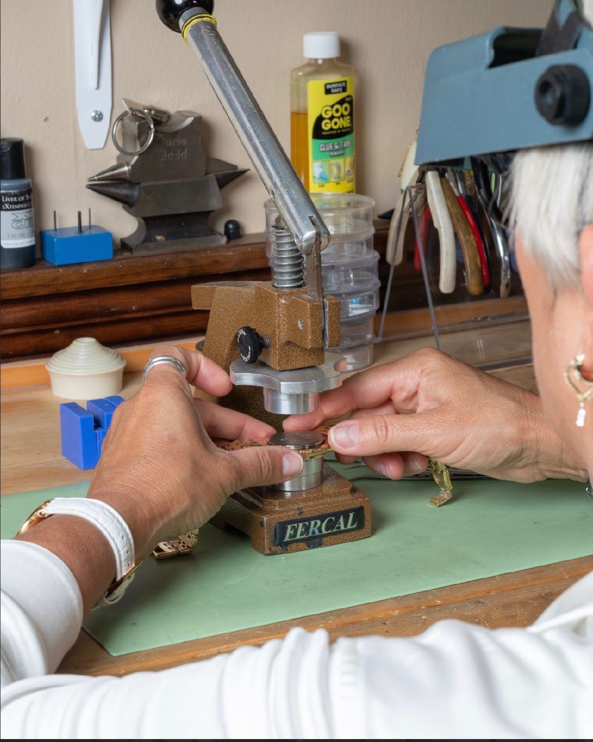 Hands using a bench-mounted press on a workbench in a workshop, with tools and supplies nearby