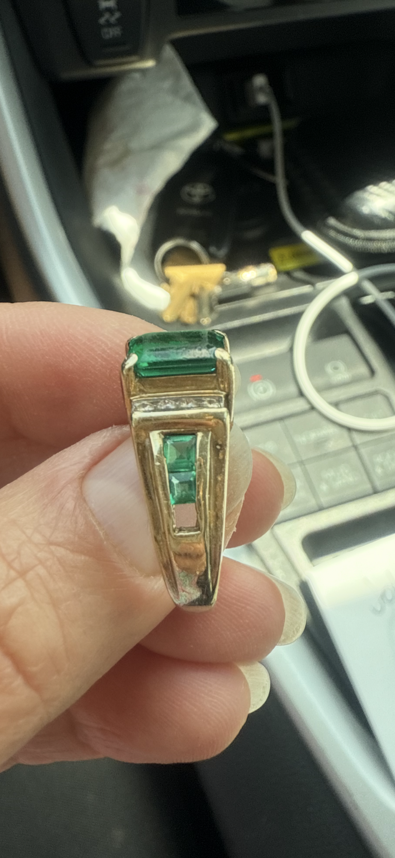 Hand holding a gold ring with green rectangular stones inside a car.