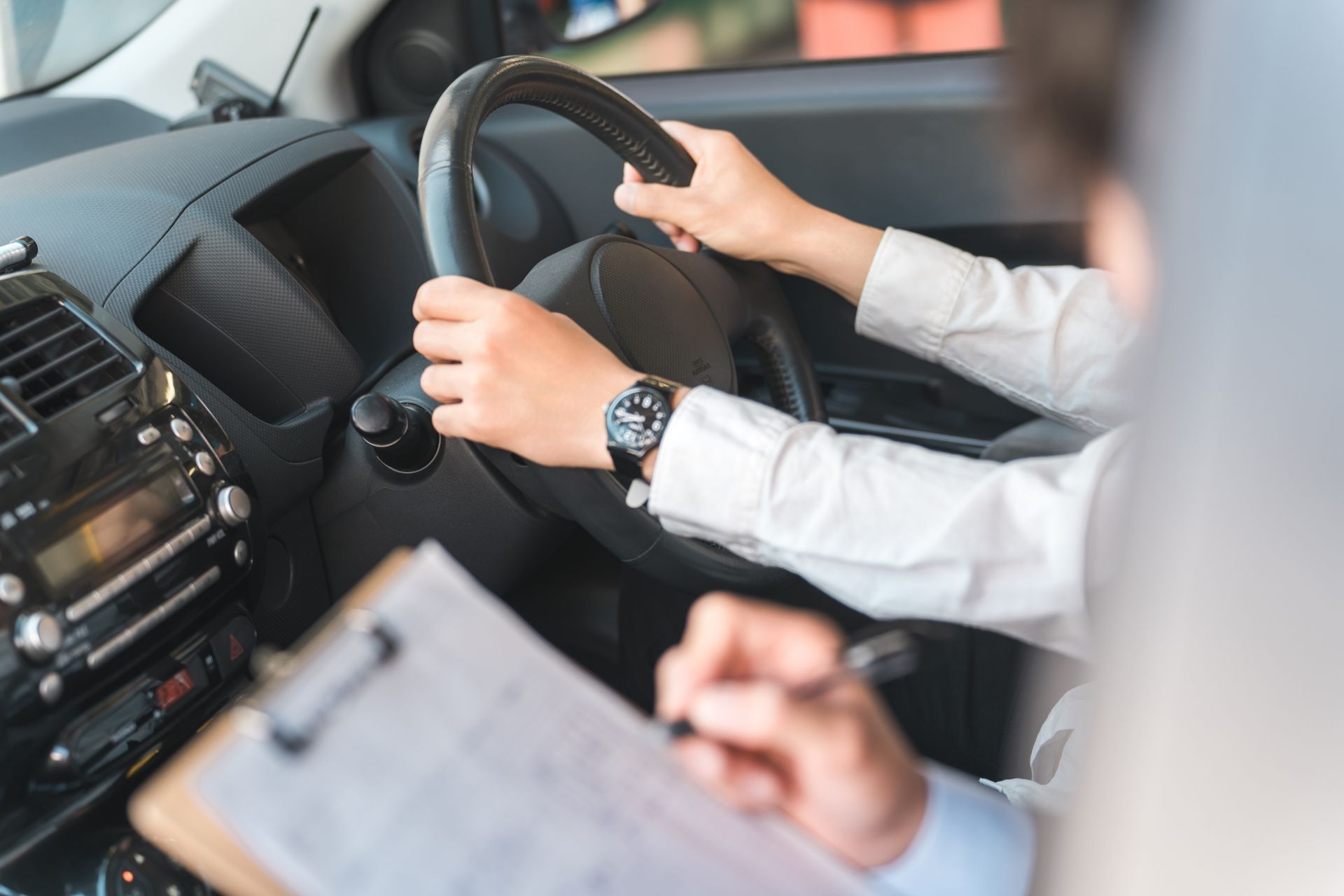 Student grips steering wheel while instructor takes notes on a clipboard during driving test.