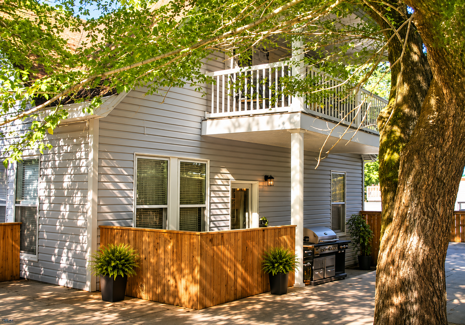 Two-story gray house with a wooden fence and balcony, shaded by a large tree on a sunny street.