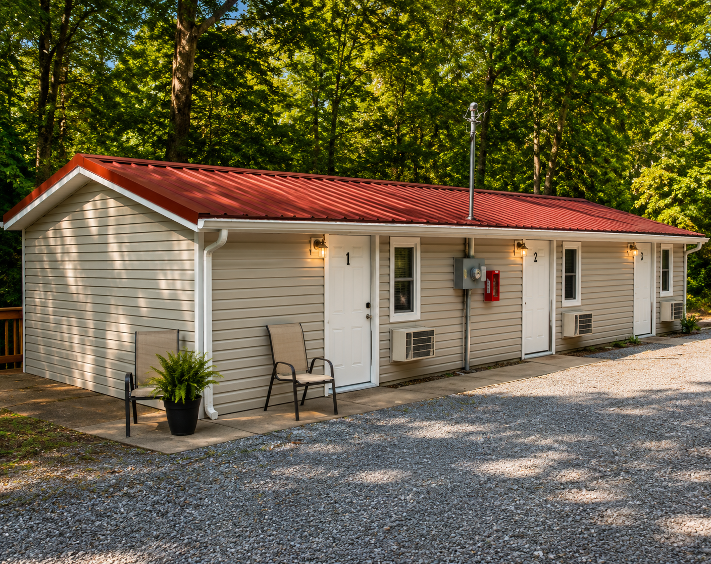 Small beige cottage with red roof, gravel driveway, and two chairs on the front porch amid trees