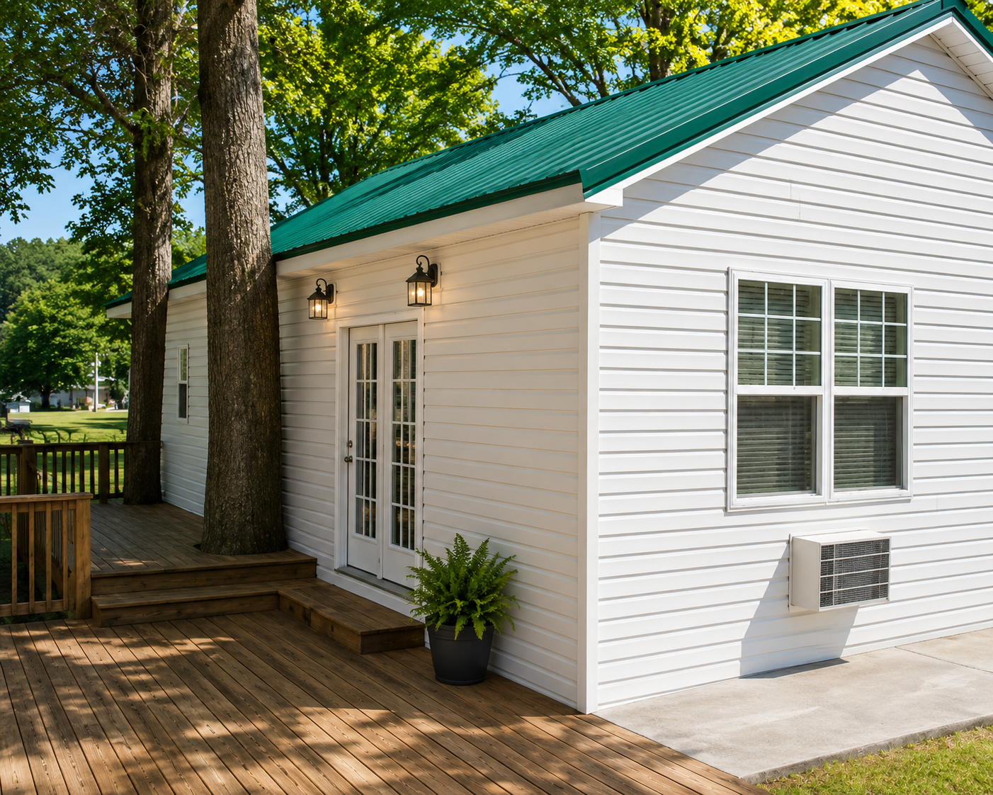 White shed with green roof and double doors on a wooden deck beside trees and a fence
