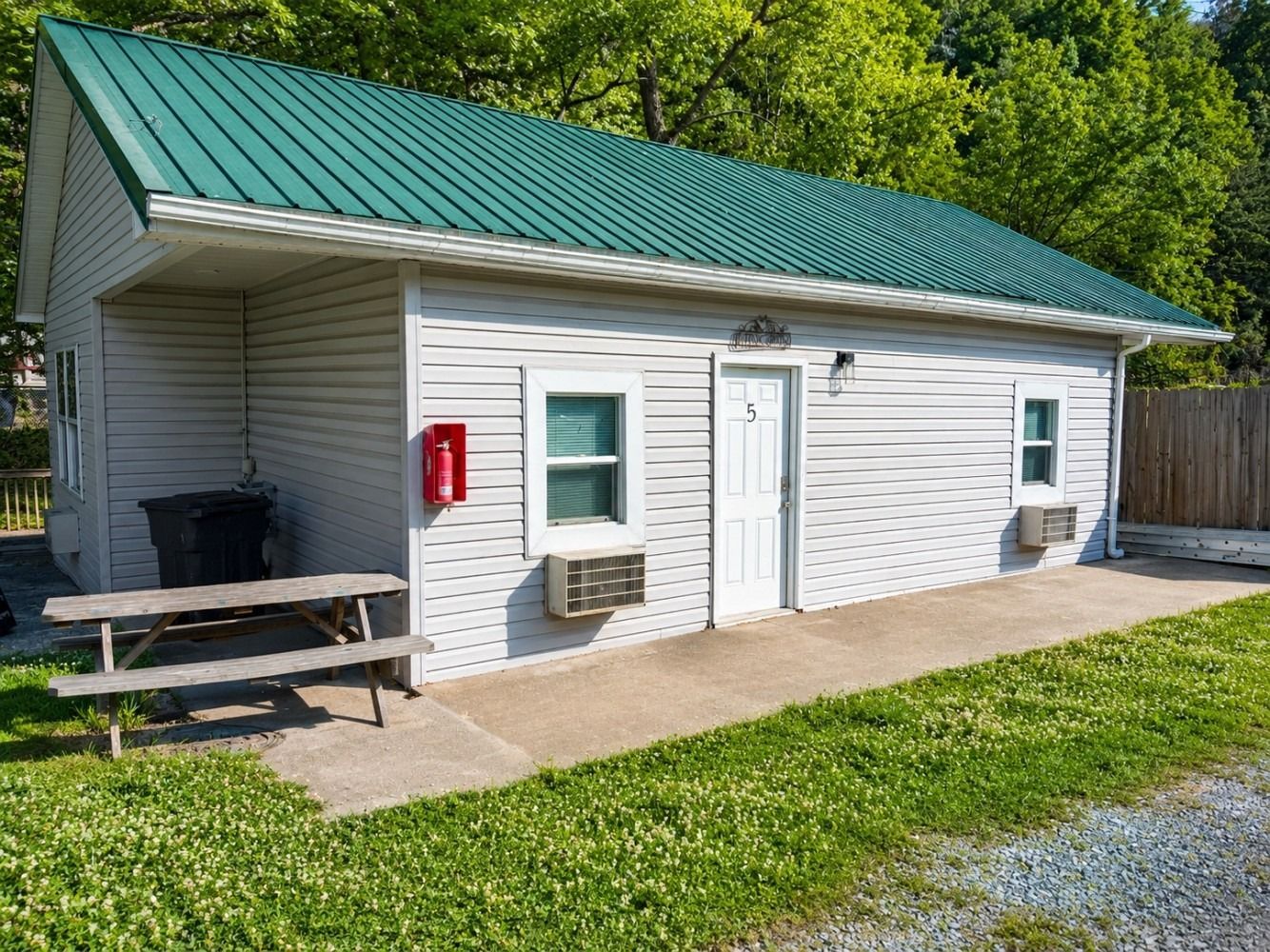Small white utility building with green roof, side door, window, and picnic table beside a grassy area.