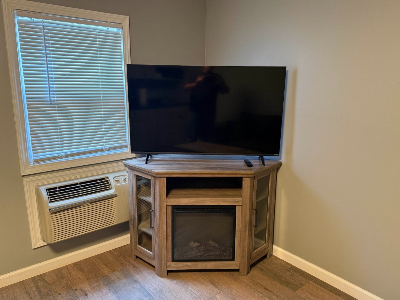 Corner living room with TV on wooden stand, window with blinds, and a wall heater underneath