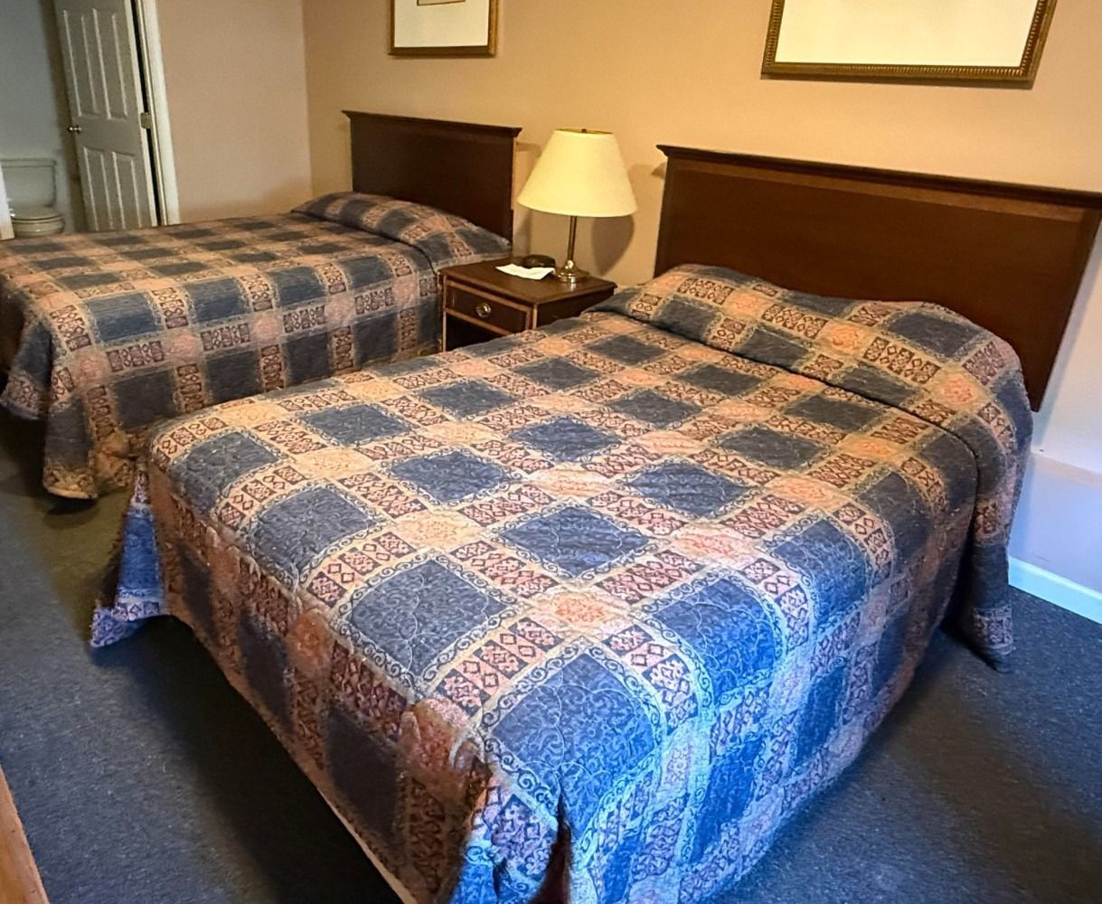 Hotel room with two unmade beds covered in blue and brown plaid quilts, a nightstand, and a lamp