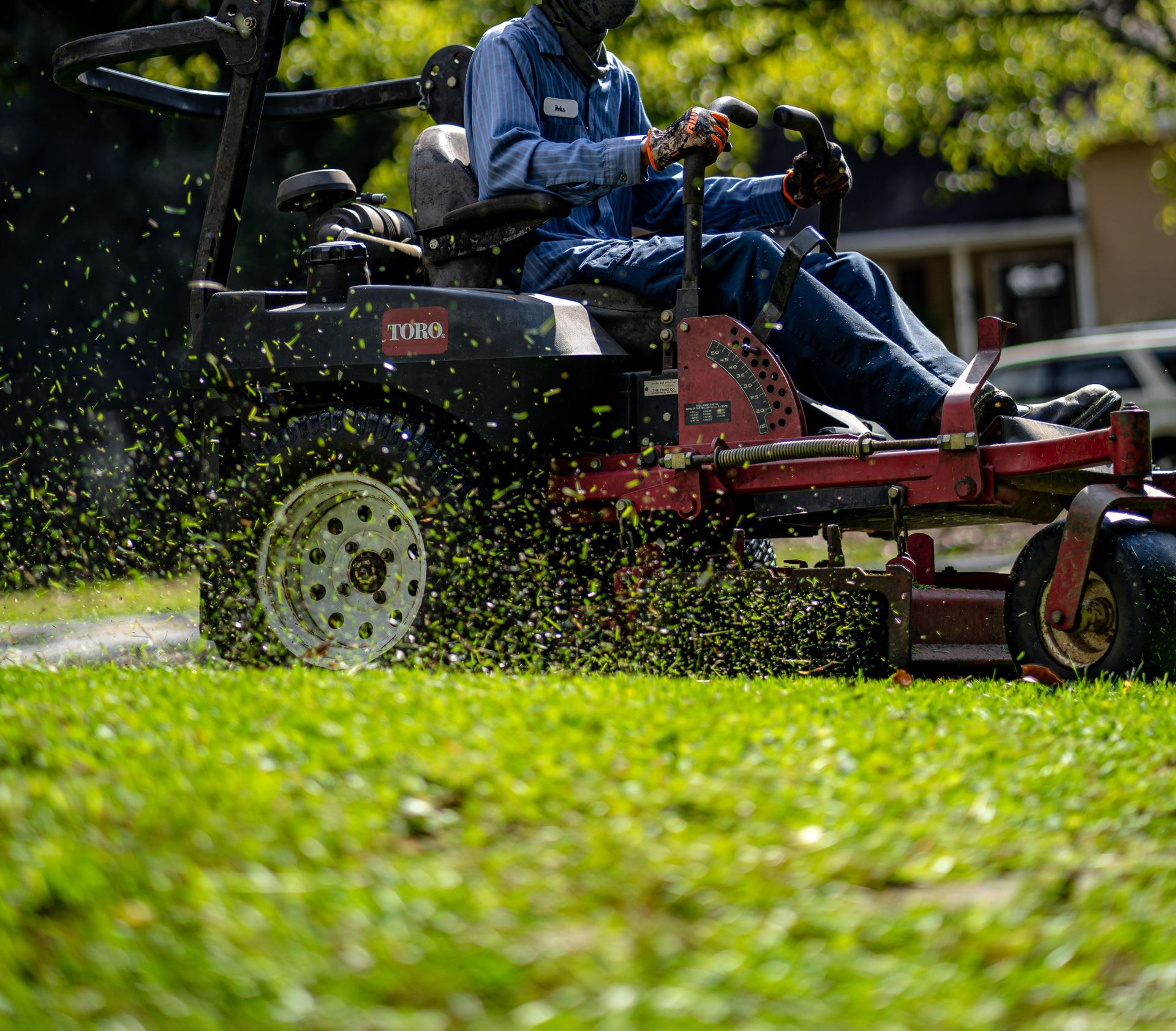 Person mowing a green lawn with a red riding mower, grass clippings flying in the sunlight.