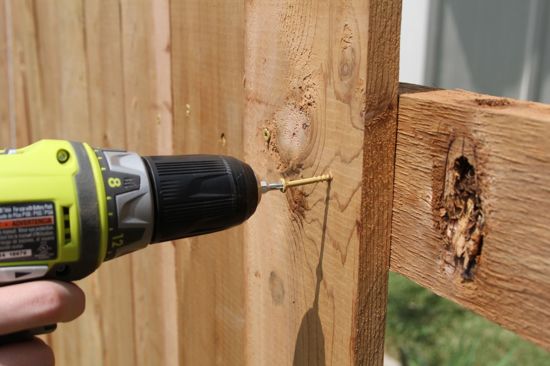 A hand drills a screw into a wooden fence post with a green power drill.