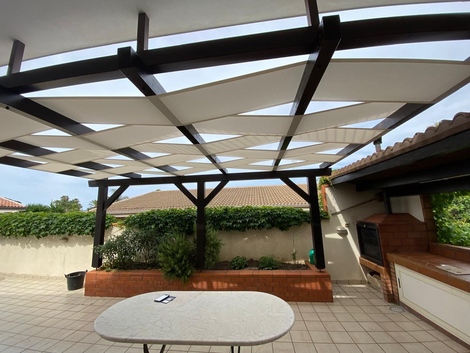 Patio con pergolato in legno e pannelli in tessuto bianco, tavolo, fioriera in mattoni e griglia.