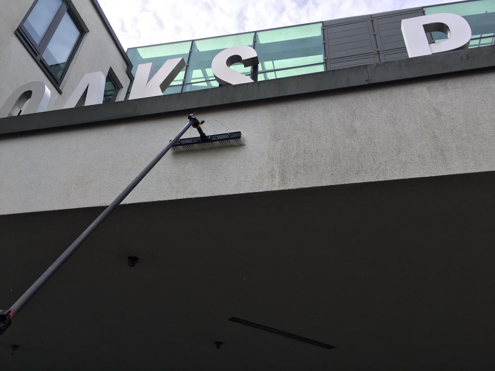 A long-handled brush cleaning a building's white exterior wall with large white letters CMKCD visible above.