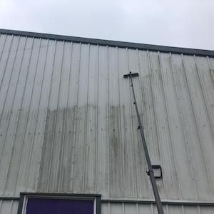 A person cleaning the exterior of a metal building with a long brush, overcast sky.