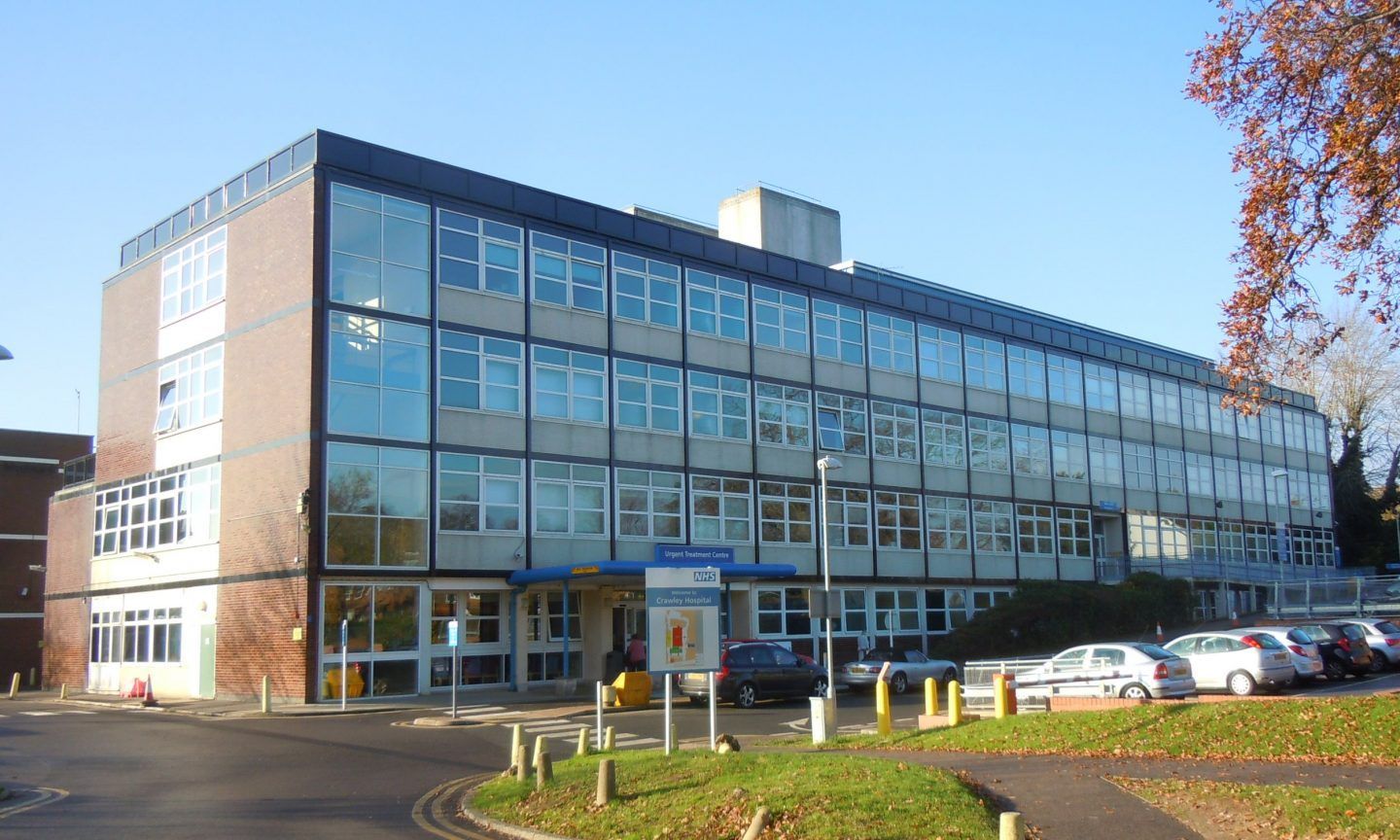 Modern, multi-story building with blue-tinted windows. Cars parked nearby. Sunny day.