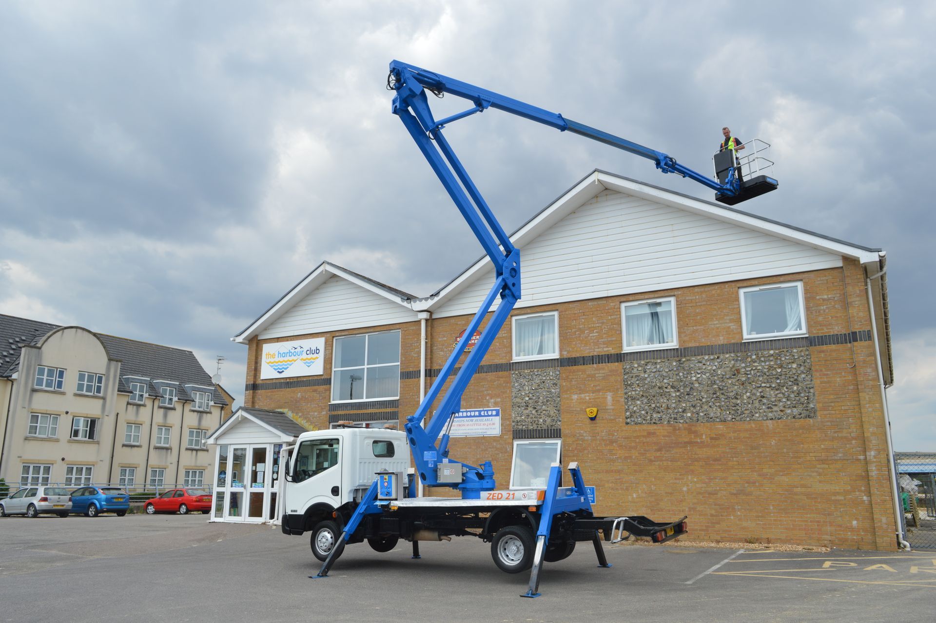 Blue cherry picker truck with extended arm reaching toward a building. Person in basket. Cloudy sky.