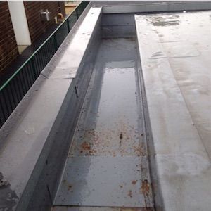 A flat, metal rooftop with a water-filled gutter. Rust spots visible.