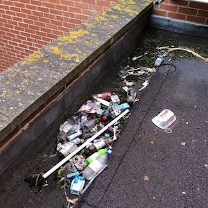 Pile of trash including bottles and containers on a flat, dirty rooftop next to a brick wall.