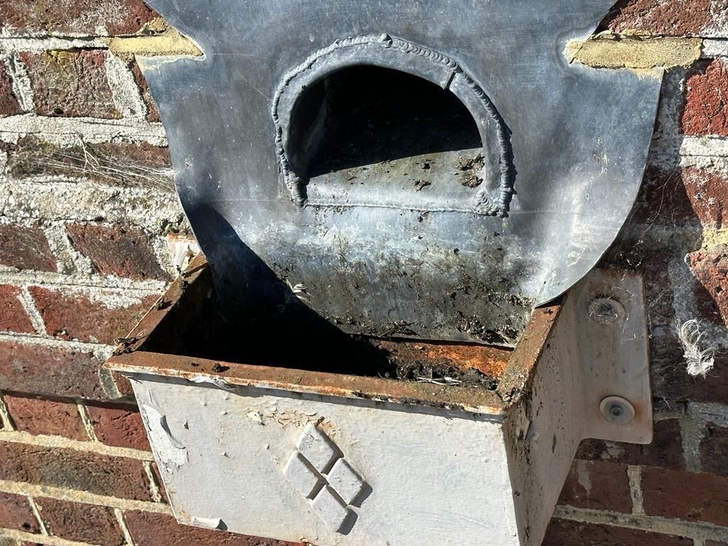 Metal birdhouse on a brick wall. The dark gray front has a round opening above a light-colored box.