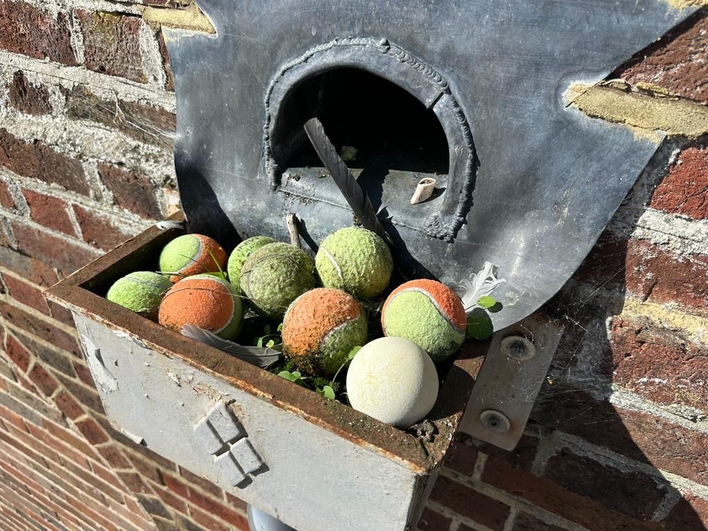Metal tennis ball dispenser on brick wall, filled with used tennis balls.