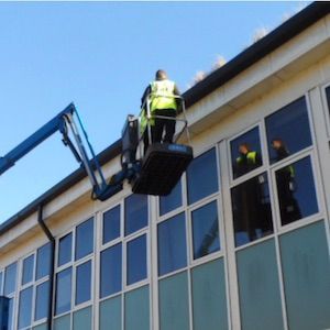Worker in lift platform, repairing roof on a building with large windows, bright sky.