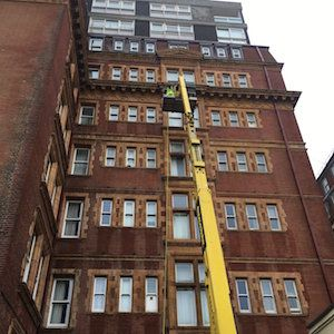 Worker in a yellow lift repairing a brick building with many windows; overcast sky.