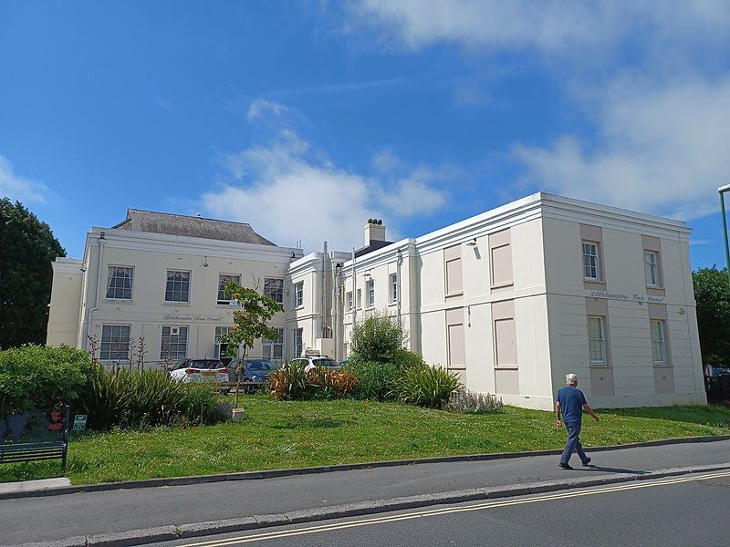 Exterior of a light-colored building with a green lawn, blue sky, and a person walking on the sidewalk.